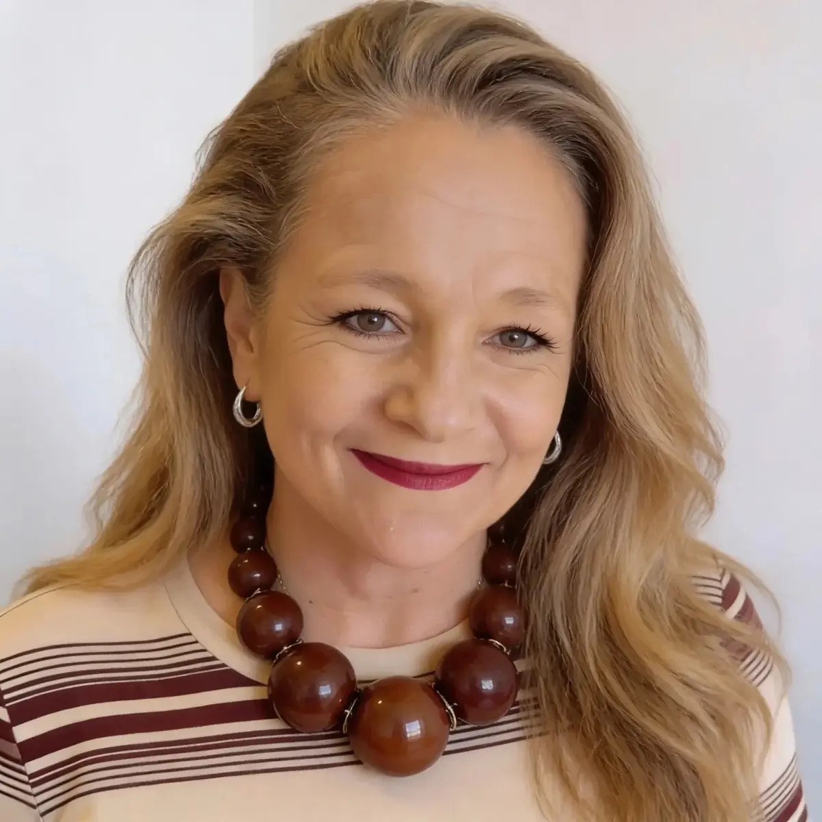 Woman with wavy blonde hair smiling, wearing a chunky brown bead necklace and striped cream and maroon top.